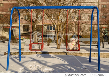 children's swing with a toy bear in the snow in the courtyard of a house children's swing with a toy bear in the snow in the courtyard of a house 135028029
