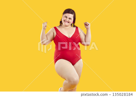 Happy joyful plus size female model in red swimsuit standing on yellow background Happy joyful plus size female model in red swimsuit standing on yellow background 135028184