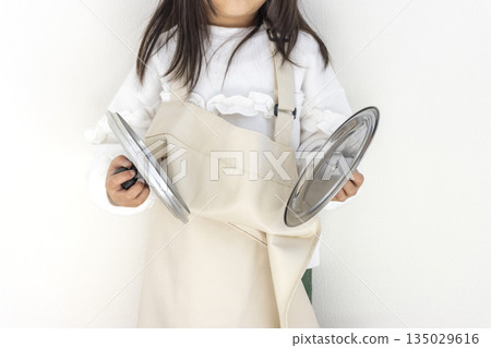 5歲日本女孩,穿著寬鬆的大圍裙,正在用鍋蓋發出聲音。 5歲日本女孩,穿著寬鬆的大圍裙,正在用鍋蓋發出聲音。 135029616