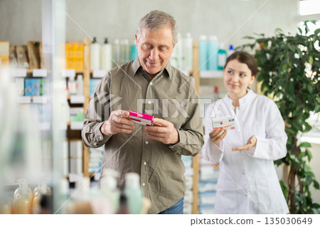 Senior man buys pills at pharmacy. Girl pharmacist in background Senior man buys pills at pharmacy. Girl pharmacist in background 135030649