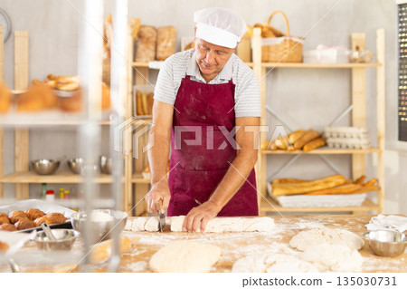 Senior man works in bakery as baker, cuts dough into portions, forms pieces of dough to create buns. 135030731
