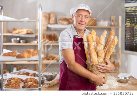 Successful aged baker holding wicker basket of baguettes in bakery Successful aged baker holding wicker basket of baguettes in bakery 135030732