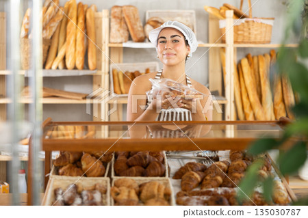 Young saleswoman holds out croissants in plastic bag in bakery Young saleswoman holds out croissants in plastic bag in bakery 135030787