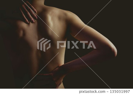 Woman's bare back and arms being lit by directional lighting in moody portrait on dark backdrop Woman's bare back and arms being lit by directional lighting in moody portrait on dark backdrop 135031997