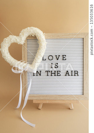 Letter board with text LOVE IS IN THE AIR Heart made of white lilac flowers on pastel beige background. Still life romantic composition of Valentines day holiday copy space for your text invitation Letter board with text LOVE IS IN THE AIR Heart made of white lilac flowers on pastel beige background. Still life romantic composition of Valentines day holiday copy space for your text invitation 135033616