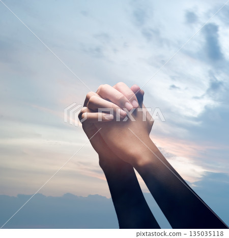 Closeup image of hands clasped together in prayer against a backdrop of a peaceful and soft colored sky. Concept of faith, hope, and spirituality 135035118
