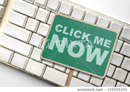 Closeup top view of a green sign with white text "Click Me Now" placed on top of a white computer keyboard. Concept of online action, call to action, and technology 135035203