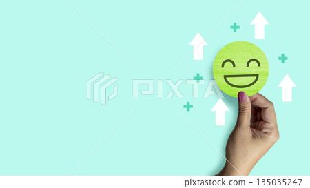 Woman hand holding a green smiling face paper circle with white arrows and a green plus sign around, on a light green background. Concept of Mental Health 135035247