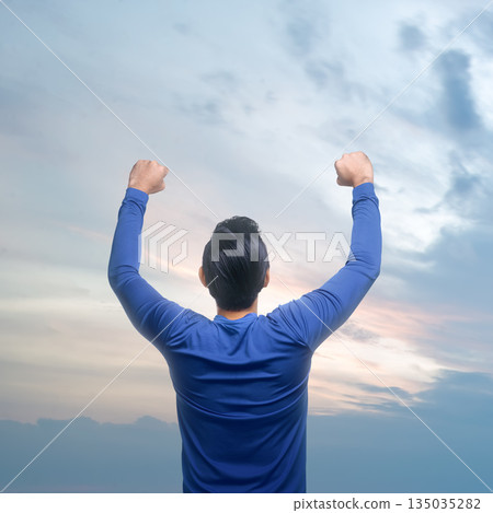 Rear view of man from the back with his arms raised up in front of a cloudscape at dusk, a feeling of freedom and achievement Rear view of man from the back with his arms raised up in front of a cloudscape at dusk, a feeling of freedom and achievement 135035282