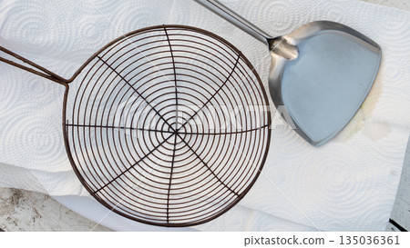Top view of wire mesh strainer and stainless steel spatula on white paper towel. Kitchen utensils for frying and draining oil concept. 135036361
