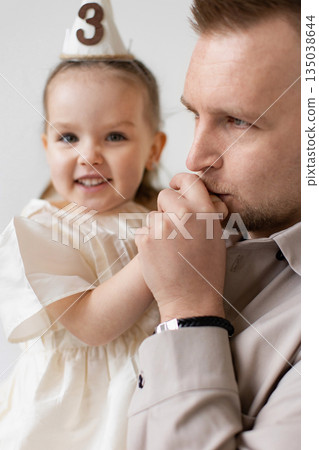 A father holds his daughter, who is wearing a birthday hat with the number 3 on it, celebrating her birthday. 135038644
