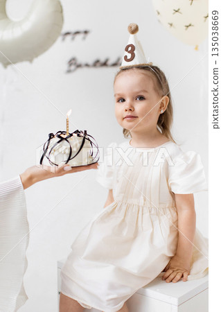 A young girl celebrates her third birthday, holding a cake with a lit candle, wearing a party hat, and sitting on a white surface. 135038669