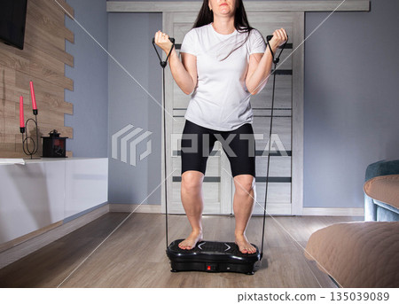 Brunette girl exercises with expanders at home on a vibration platform to reduce weight and cellulite. Exercise at home, lifestyle 135039089