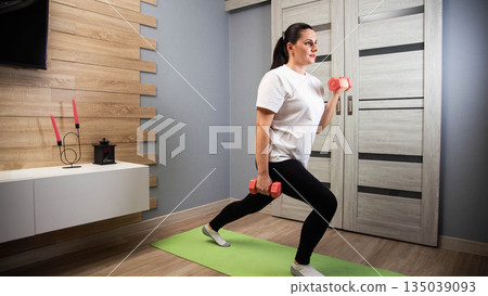 Woman performs lunge exercise with dumbbells indoors, fitness training for physical strength and well-being, healthy lifestyle at home 135039093