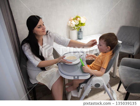 Caring woman feeds young boy sitting in high chair indoors, showing tender moment of mealtime between parent and child at home Caring woman feeds young boy sitting in high chair indoors, showing tender moment of mealtime between parent and child at home 135039135