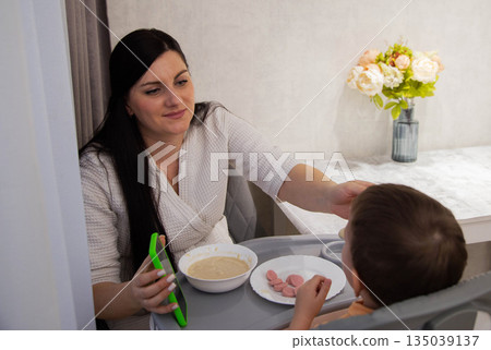 Woman feeding baby in high chair indoors with porridge and sausages. Mother child interaction during breakfast. Daily routine of parent and infant. Woman feeding baby in high chair indoors with porridge and sausages. Mother child interaction during breakfast. Daily routine of parent and infant. 135039137