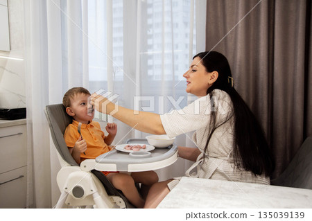 Woman feeding young child in high chair, child wears orange polo shirt, food on face, domestic scene of care and mealtime Woman feeding young child in high chair, child wears orange polo shirt, food on face, domestic scene of care and mealtime 135039139