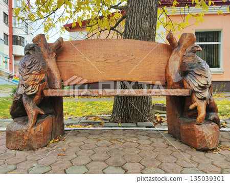 Elaborate handcrafted carved wooden bench featuring animal figure supports, such as owls or eagles, nestled in public park setting amidst fallen autumn leaves 135039301