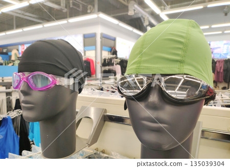 Mannequin heads displaying swimming caps and goggles in retail store, showcasing sport equipment for water activity, selection of protective eyewear and headwear for swimmers 135039304