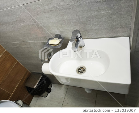 White ceramic wash basin with chrome faucet and bar soap in clean bathroom interior gray wall tiles and brown floor tiles for hygiene and cleanliness 135039307