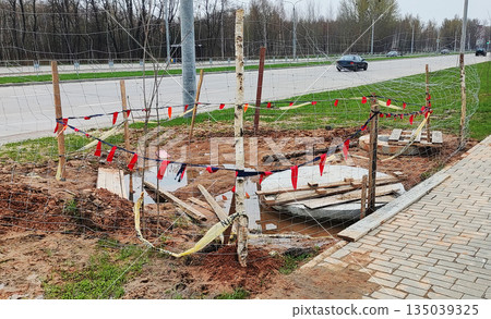 Construction site alongside urban road, temporary fencing with caution tape and red flags securing excavation area, visible debris and standing water, vehicle traffic passing by, overcast day. 135039325