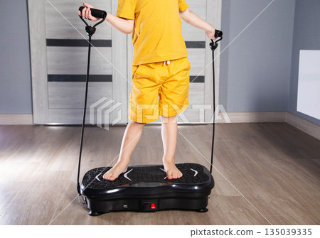 Handsome 3 year old boy in yellow suit sits on modern vibration platform for exercising at home, background 135039335