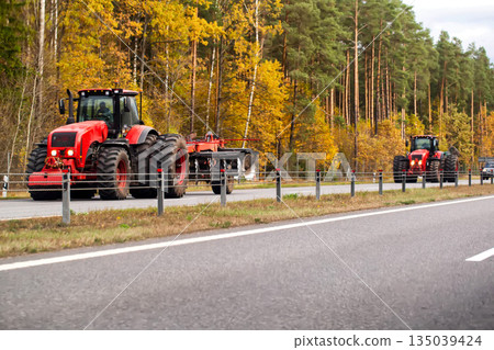 Red tractors driving on highway against background of autumn forest with vibrant yellow foliage and tall green pine trees, agricultural machinery transport on rural road Red tractors driving on highway against background of autumn forest with vibrant yellow foliage and tall green pine trees, agricultural machinery transport on rural road 135039424