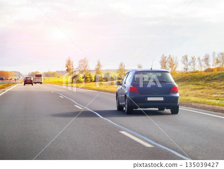 Blue hatchback car driving on highway during autumn day. Road trip scene with vehicle traveling at speed. Transportation concept with bright sun flare. 135039427