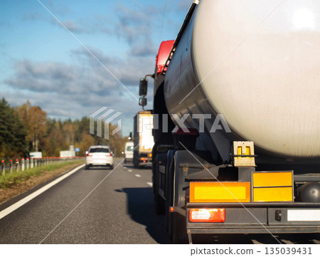 Rear view of white tanker truck driving on multilane highway, showing back lights and orange reflector plates, with other vehicles and autumn trees along road under partly cloudy blue sky. Rear view of white tanker truck driving on multilane highway, showing back lights and orange reflector plates, with other vehicles and autumn trees along road under partly cloudy blue sky. 135039431