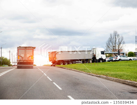 Long haul truck on highway; another truck with trailer parked roadside; freight transportation scene with industrial background under cloudy sky Long haul truck on highway; another truck with trailer parked roadside; freight transportation scene with industrial background under cloudy sky 135039434