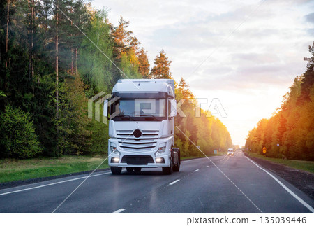 White semi-trailer truck drives on paved rural road through dense forest. Sunlight illuminates trees during sunset. Essential freight transportation. White semi-trailer truck drives on paved rural road through dense forest. Sunlight illuminates trees during sunset. Essential freight transportation. 135039446