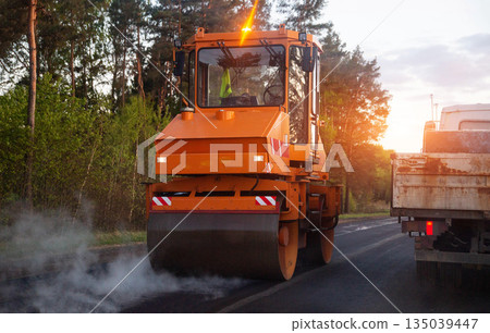 Orange road roller compacting hot asphalt on rural road during construction project, steam rising from new pavement under sunset light 135039447