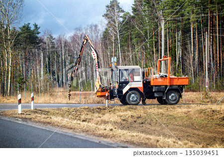 An orange and white utility vehicle with a drilling boom is parked on the side of a paved road during daylight hours. Road maintenance and pole installation. An orange and white utility vehicle with a drilling boom is parked on the side of a paved road during daylight hours. Road maintenance and pole installation. 135039451