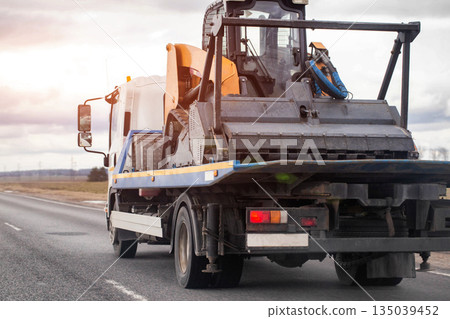 Heavy duty truck transporting compact track loader on open road under cloudy sky, showing rear view of industrial vehicle and specialized cargo 135039452