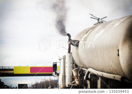 Large industrial truck traveling on highway with thick black exhaust smoke rising from tall smokestack, visible against overcast sky. Copy space for text Large industrial truck traveling on highway with thick black exhaust smoke rising from tall smokestack, visible against overcast sky. Copy space for text 135039453