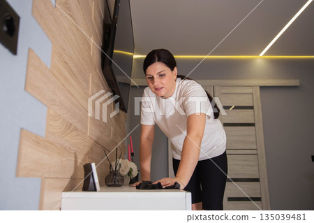 Woman wiping dust from surface of white furniture in modern apartment living room, home cleaning routine, domestic chore, daily dwelling maintenance. Woman wiping dust from surface of white furniture in modern apartment living room, home cleaning routine, domestic chore, daily dwelling maintenance. 135039481