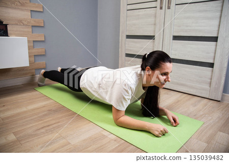 Woman performing plank exercise on yoga mat indoors. Fitness routine in apartment. Core strength workout. Healthy lifestyle activity for adults. Woman performing plank exercise on yoga mat indoors. Fitness routine in apartment. Core strength workout. Healthy lifestyle activity for adults. 135039482