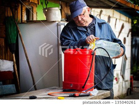 Elderly man prepares solution for plant pest control, pouring liquid from plastic bottle into red garden sprayer for dacha agriculture work 135040261