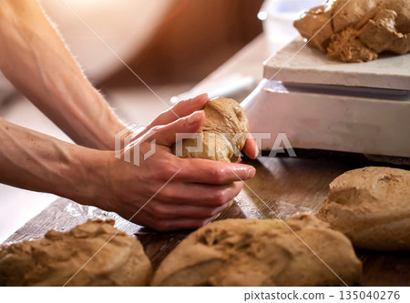 Hands shaping dough for baking preparation in kitchen. Close up of baker working with raw bread dough on rustic wooden table, illuminated by warm light, creating artisan food product. Hands shaping dough for baking preparation in kitchen. Close up of baker working with raw bread dough on rustic wooden table, illuminated by warm light, creating artisan food product. 135040276