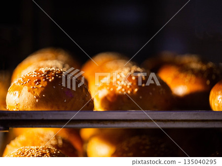 Golden-brown bread buns with sesame seed topping cooling on metal rack in bakery environment during manufacturing process, fresh baked goods display 135040295