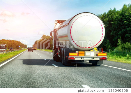 White tank truck with flammable cargo transports fuel on multi-lane asphalt highway through green forest area on clear day. Copy space for text White tank truck with flammable cargo transports fuel on multi-lane asphalt highway through green forest area on clear day. Copy space for text 135040306