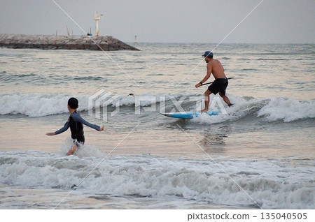 A young surfer rides a wave on a wooden board in the blue sea. A child plays in the waves nearby.Young surfer and child. A young surfer rides a wave on a wooden board in the blue sea. A child plays in the waves nearby.Young surfer and child. 135040505