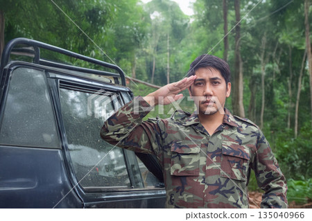 An Asian army officer in uniform salutes directly at the camera, deep in a lush jungle with an off-road vehicle visible in the background 135040966