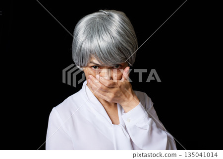 A white-haired senior woman standing in the middle of a black background, covering her mouth with her hand and glaring A white-haired senior woman standing in the middle of a black background, covering her mouth with her hand and glaring 135041014