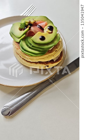 Stacked pancake with avocado and salmon served on plate with sauce and cutlery on white background. This breakfast food represents healthy lifestyle and balanced gourmet choice Stacked pancake with avocado and salmon served on plate with sauce and cutlery on white background. This breakfast food represents healthy lifestyle and balanced gourmet choice 135041947
