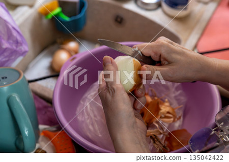 Close-Up Of Hand Peeling An Onion With Knife Over Purple Bowl In The Kitchen Close-Up Of Hand Peeling An Onion With Knife Over Purple Bowl In The Kitchen 135042422
