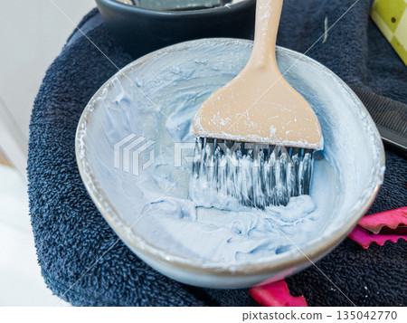 Rich, creamy hair dye mix rests on a wooden brush over a sleek bowl. The tools for hair coloring lie on a dark, textured towel, with a hint of vibrant pink peeking through, suggesting the lively work 135042770