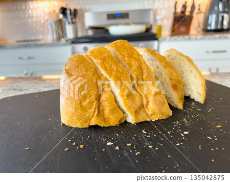 Loaf of freshly baked bread sliced into pieces and arranged on a black cutting board in a home kitchen. 135042875