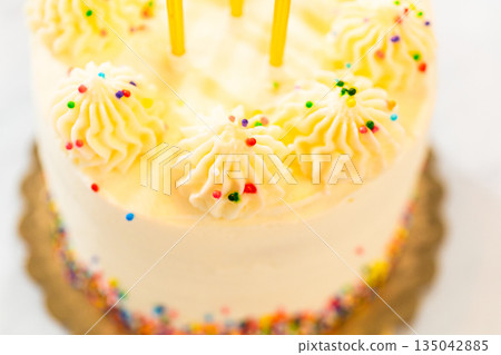A close side view of a birthday cake topped with yellow candles and swirled frosting, placed on a gold-colored base. The frosting is decorated with small colorful sprinkles, adding vibrant detail. 135042885