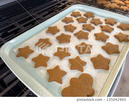 A baking tray lined with parchment paper holds freshly baked gingerbread cookies in star and gingerbread-person shapes. The tray rests on a stovetop with warm kitchen lighting revealing their golden 135042903